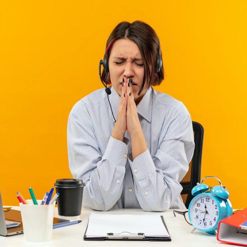 anxious young call center girl wearing headset sitting at desk putting hands in pray gesture with closed eyes isolated on orange background